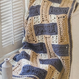 May include: A handmade crochet blanket in a geometric pattern of navy blue, beige, and cream. The blanket is draped over a windowsill, with a small vase in the foreground. The text in the image says, "Here's a throw that's easy enough for a beginner, yet offers out-of-the-ordinary style."