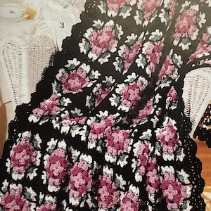 May include: A black crocheted blanket featuring a floral design in pink, white, and green. The blanket is draped over a white wicker chair and has a black scalloped border.