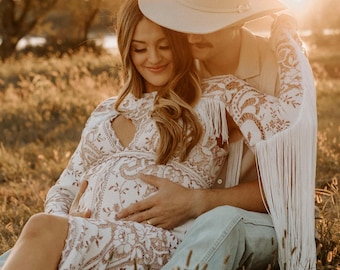 May include: A pregnant woman in a white lace dress with a fringe trim sits in a field with a man wearing a cowboy hat. The man has his arm around the woman and is holding her belly.