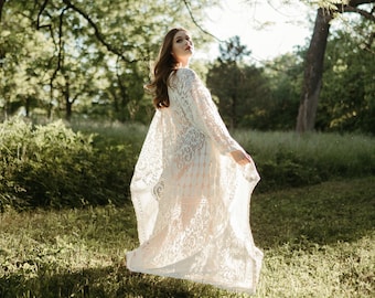 May include: A woman wearing a white lace maxi dress with long sleeves and a flowing skirt. She is standing in a grassy field with trees in the background.