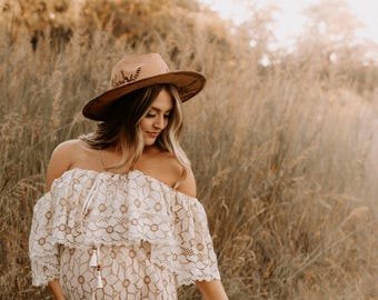 May include: A pregnant woman wearing a white off-the-shoulder lace dress with a brown hat. She is standing in a field of tall grass.