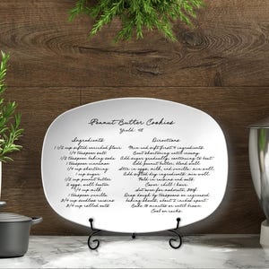 May include: A white oval ceramic platter featuring black printed text of a Peanut Butter Cookies recipe. The text includes a yield of 48, a list of ingredients, and step-by-step directions in a cursive script font, displayed on a black metal stand.