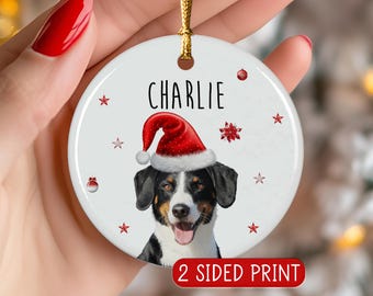 May include: A round, white ceramic Christmas ornament featuring a dog wearing a Santa hat. The dog's name, "Charlie," is printed above the image. Red stars and snowflakes decorate the background. The text "2 SIDED PRINT" is at the bottom.