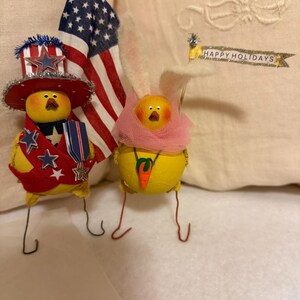 Peut inclure: Deux figurines de poussins jaunes, l'une vêtue d'une tenue patriotique d'Oncle Sam avec un chapeau rouge, blanc et bleu, l'autre avec des oreilles de lapin et une écharpe rose. Un drapeau américain est en arrière-plan, et une bannière indique "HAPPY HOLIDAYS."
