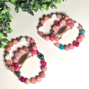 May include: Two beaded bracelets with a variety of colorful gemstones. The bracelets feature pink, white, and green beads, with a central white, oval-shaped accent. Rose gold-colored metal beads are interspersed throughout.