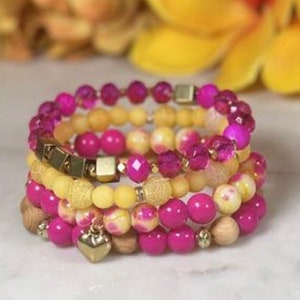 Peut inclure: Plusieurs bracelets empilés, aux tons rose, jaune et or. Les bracelets sont composés de différents types de perles, dont des perles facettées, rondes et en bois. Un bracelet est orné d'un charm en forme de cœur doré.