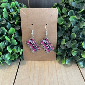 May include: A pair of dangle earrings featuring a cassette tape design. The earrings have a clear background with a pink and gray cassette tape graphic. The earrings are displayed on a brown card with a wooden background.