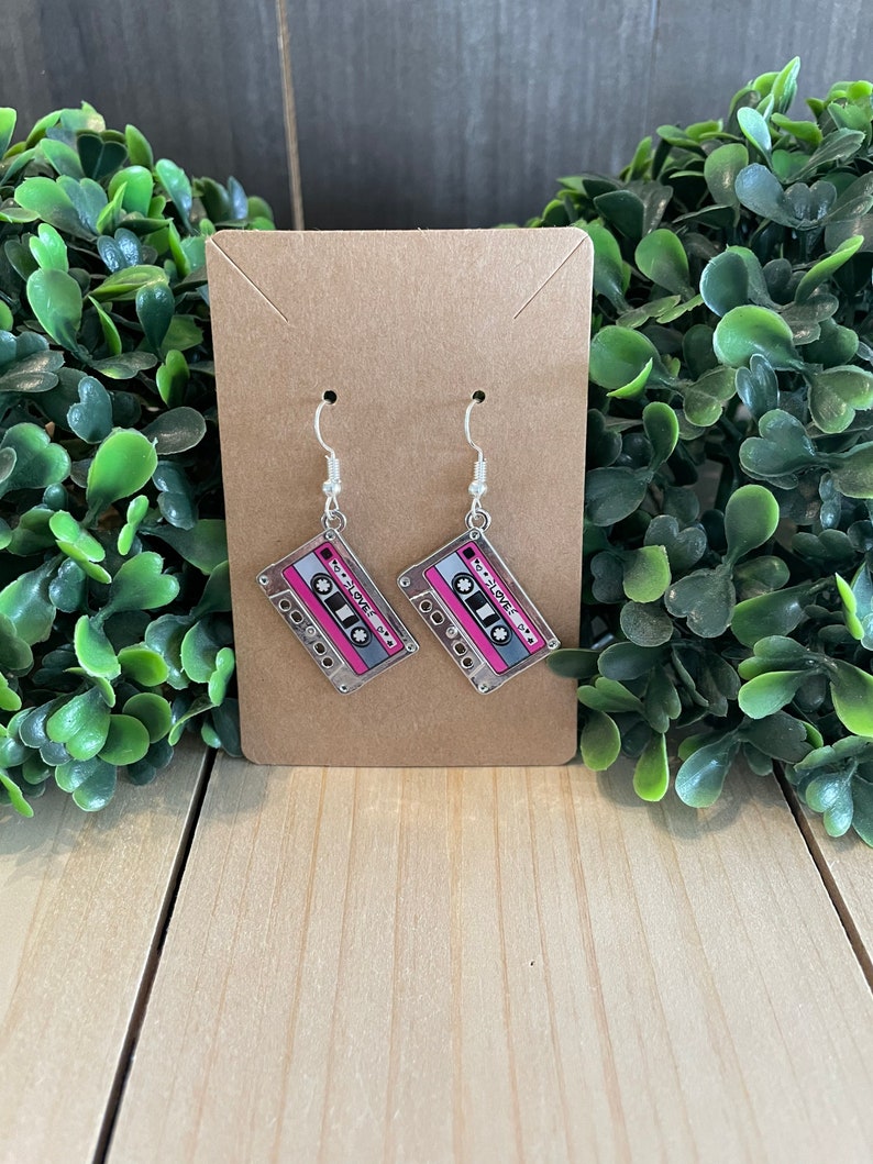 May include: A pair of dangle earrings featuring a cassette tape design. The earrings have a clear background with a pink and gray cassette tape graphic. The earrings are displayed on a brown card with a wooden background.