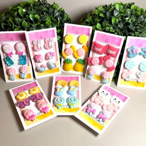 May include: Assortment of colorful, themed earring sets on pink and yellow cards.  The sets include candy, pineapple, flowers, butterflies, and cartoon characters.  The cards have the text "Sweet & Stylish" printed on them.