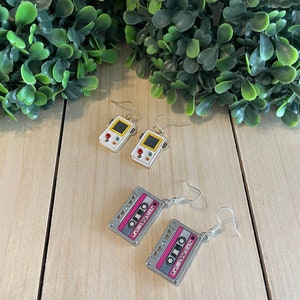May include: Earrings featuring retro designs. One pair is shaped like a handheld video game console with a yellow screen and red buttons. The other pair is a cassette tape with pink accents and the text "I LOVE". Both pairs have silver-colored hooks.