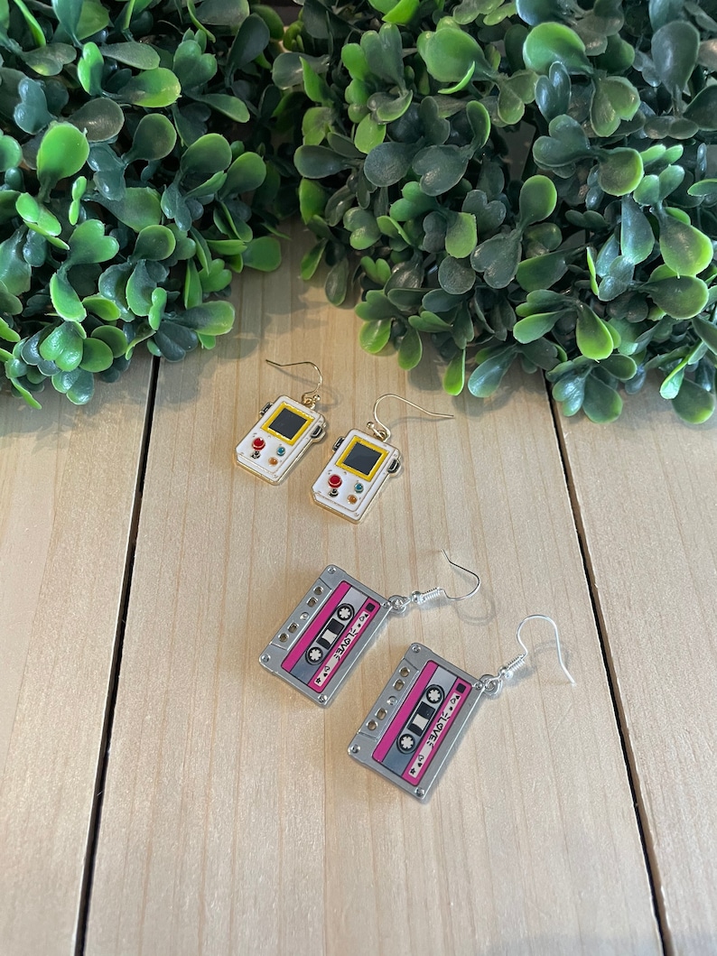 May include: Earrings featuring retro designs. One pair is shaped like a handheld video game console with a yellow screen and red buttons. The other pair is a cassette tape with pink accents and the text "I LOVE". Both pairs have silver-colored hooks.