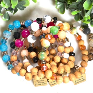 Pode incluir: Uma coleção de pulseiras de contas com uma variedade de contas coloridas, incluindo contas azuis, rosa, brancas e de madeira. Algumas pulseiras têm espaçadores dourados e pequenas etiquetas com texto. As pulseiras estão dispostas sobre uma superfície branca.
