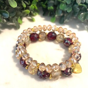 May include: Two beaded bracelets with gold accents and a gold heart charm. The bracelets feature clear, burgundy, and gold-colored beads. The bracelets are stacked together, creating a layered look. The background is a white surface with green foliage.