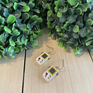 May include: White and gold-colored earrings shaped like retro handheld game consoles. Each earring features a black screen, red and blue buttons, and a yellow border. The earrings hang from gold-colored ear wires.