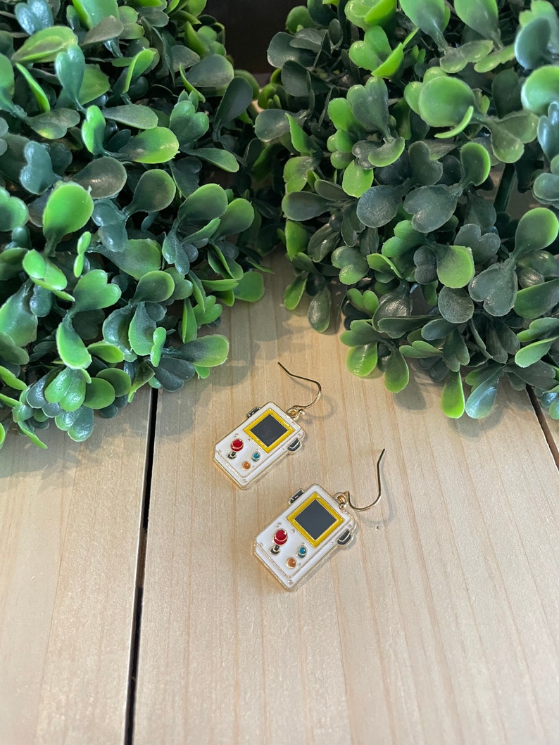 May include: White and gold-colored earrings shaped like retro handheld game consoles. Each earring features a black screen, red and blue buttons, and a yellow border. The earrings hang from gold-colored ear wires.