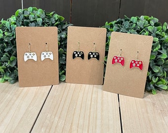 May include: Three pairs of dangle earrings featuring video game controller designs. One pair is white, one is black, and one is red. Each controller has gold-colored accents and is displayed on a brown card.