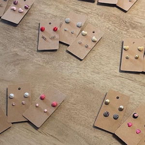Puede incluir: Surtido de juegos de pendientes mostrados en cartulina marrón. Los pendientes son de varias formas y colores, incluyendo corazones, círculos y rayas. Algunos pendientes tienen un acabado perlado.