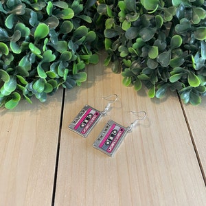 May include: A pair of silver-toned cassette tape earrings. The earrings feature a pink and gray design with the word "Love" printed on the tape. The earrings hang from silver-colored fish hook ear wires.