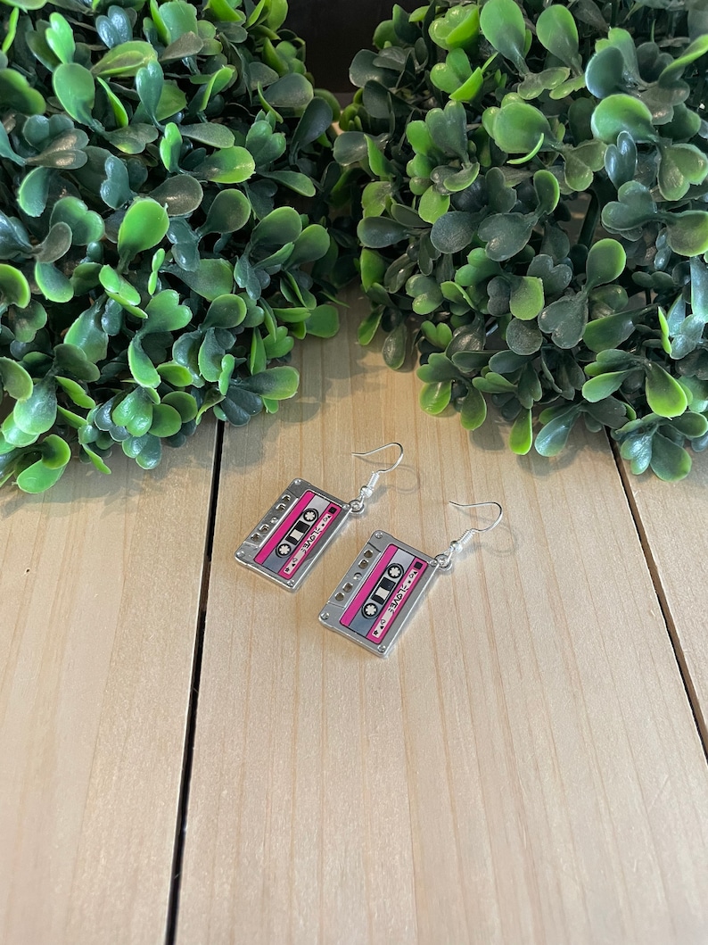 May include: A pair of silver-toned cassette tape earrings. The earrings feature a pink and gray design with the word "Love" printed on the tape. The earrings hang from silver-colored fish hook ear wires.
