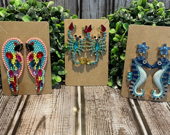 May include: Three pairs of colorful jeweled earrings displayed on brown card stock. The earrings are shaped like parrots, scorpions, and seahorses.