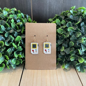 May include: A pair of white and gold earrings shaped like retro handheld game consoles. Each earring features a yellow screen, red and white buttons, and a gold hook. The earrings are displayed on a brown card.