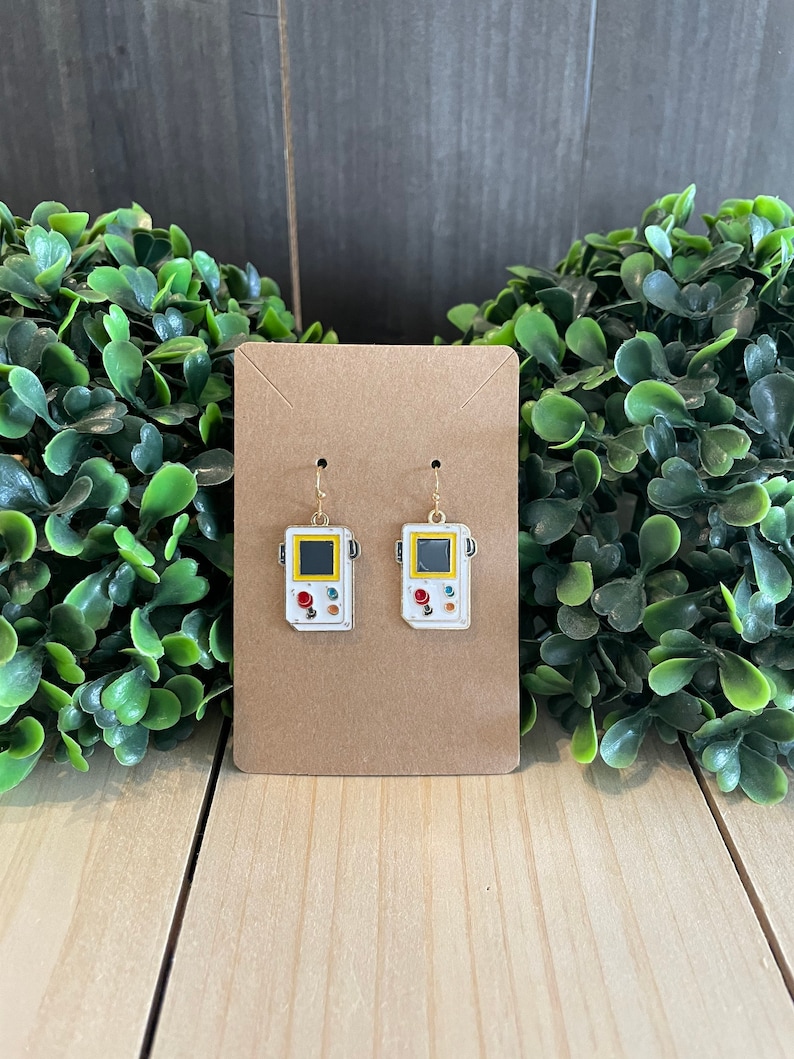 May include: A pair of white and gold earrings shaped like retro handheld game consoles. Each earring features a yellow screen, red and white buttons, and a gold hook. The earrings are displayed on a brown card.