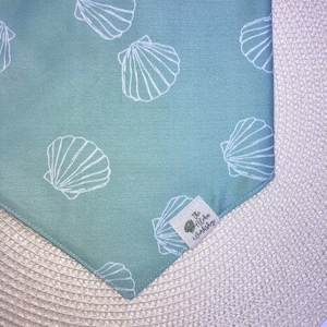 May include: A light blue bandana with a white seashell pattern. The bandana has a white label with the text "The Aloha Workshop" in black.