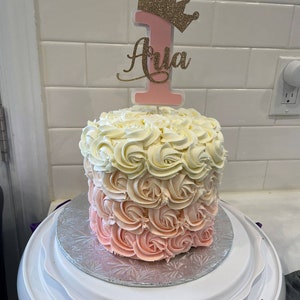May include: A pink and white ombre cake decorated with buttercream frosting in a rose pattern. The cake is topped with a pink number one and a gold glitter crown with the name "Aria" written on it.