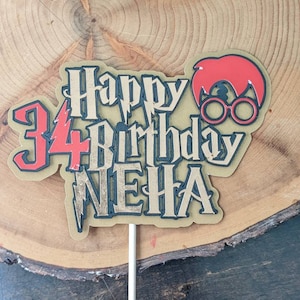 May include: A gold and black cake topper with the text "Happy 34th Birthday Neha" and a red and gold illustration of a person wearing glasses.