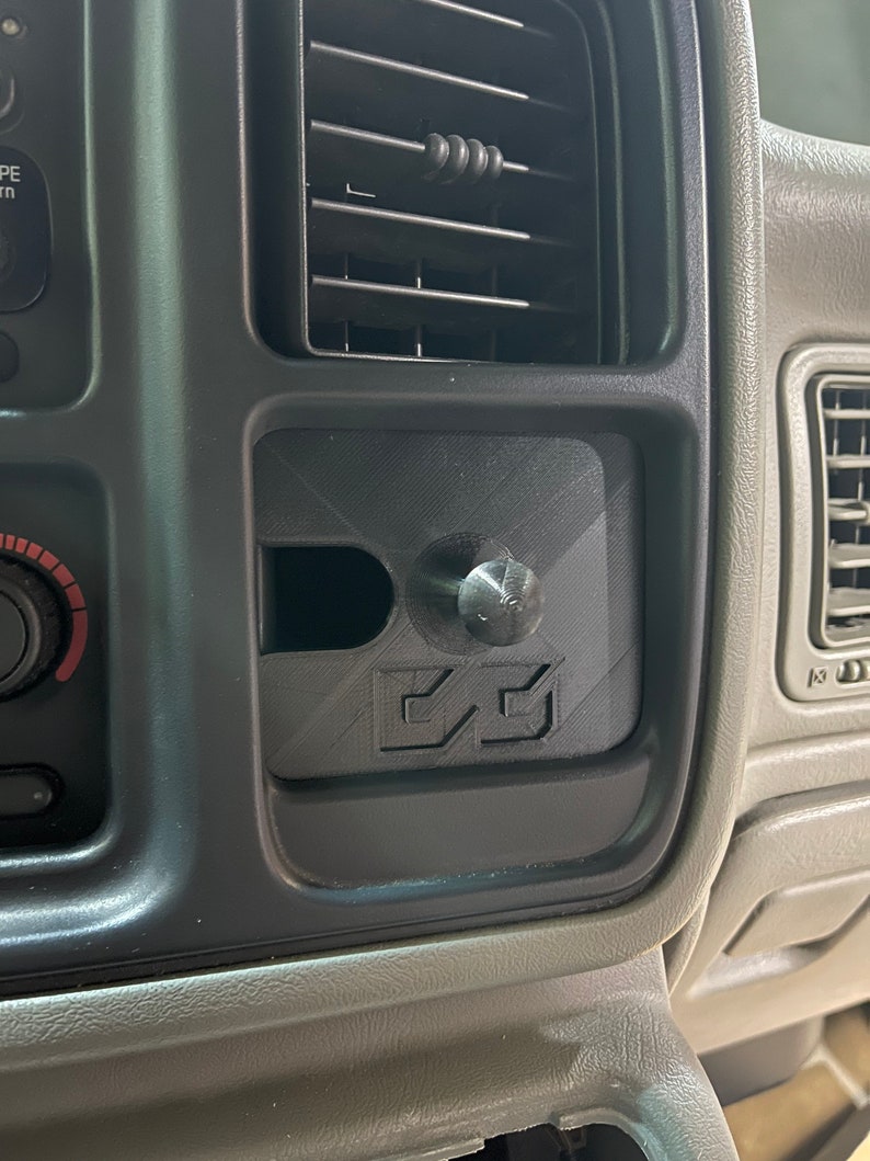 May include: A black plastic car dashboard accessory with a black knob and a 3D printed design. The design features two arrows pointing upwards.