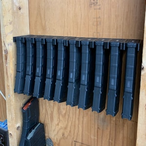 May include: A wall-mounted storage rack with 10 black magazine holders. The holders are arranged in a row and are made of a durable material.