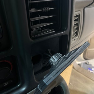 May include: A black plastic car vent mount with a knob to adjust the angle. The mount is attached to a car's air vent.