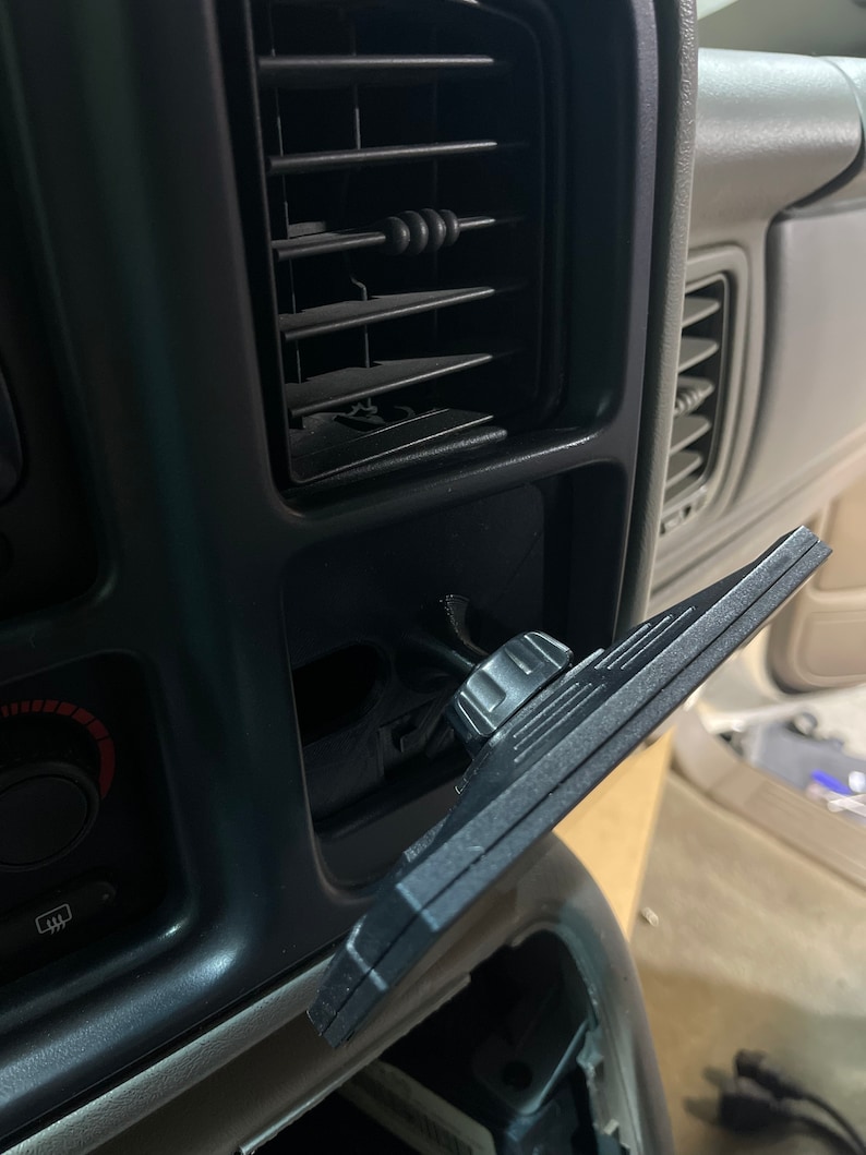 May include: A black plastic car vent mount with a knob to adjust the angle. The mount is attached to a car's air vent.