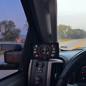 May include: A black digital gauge display mounted in a car's interior shows a variety of readings, including speed, boost, and engine temperature. The gauge is displaying a boost reading of 64.