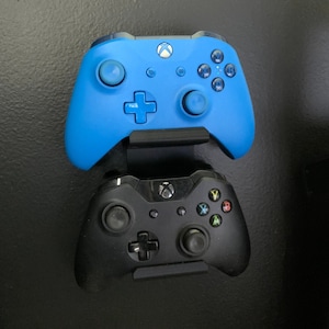 Peut inclure: Deux manettes Xbox montées sur un mur. La manette du haut est bleue et la manette du bas est noire.