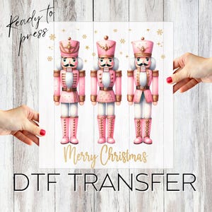 May include: Three pink and gold nutcracker figurines stand in a row on a white woodgrain background. The text "Merry Christmas" is written in gold below the figurines. The text "Ready to Press" is written in black at the top left corner of the image. The text "DTG Transfer" is written in black at the bottom of the image.