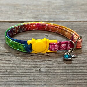 May include: A colourful cat collar with a yellow cat-shaped buckle and a blue crystal charm. The collar is made of fabric with a tie-dye pattern.