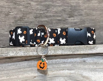 May include: Black and white dog collar with a pattern of ghosts and pumpkins. The collar has a black buckle and a silver D-ring. A silver charm with a pumpkin face is attached to the D-ring.