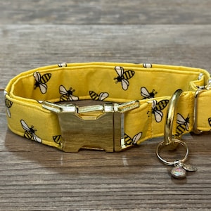 Puede incluir: Un collar de perro amarillo con un patrón de abejas y una hebilla y un anillo en D dorados. El collar tiene un amuleto dorado unido al anillo en D.