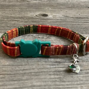 Op de afbeelding: Een rode, groene en gouden kattenhalsband met een turquoise veiligheidssluiting en een zilveren hartvormige hanger met een groene steen. De hanger heeft de tekst "Ik hou van je" erop.