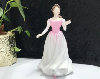 May include: A porcelain figurine of a woman in a long white dress with a pink bodice. The figurine has brown hair and is posed with one arm extended. The dress has a flowing skirt and the figurine is standing on a white surface.