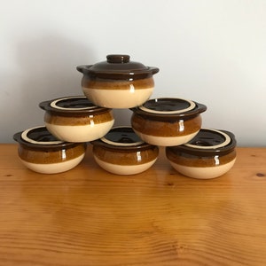 May include: Six ceramic brown and beige lidded bowls stacked in a pyramid formation. The bowls have a glossy finish and are arranged with one on top, two in the middle, and three at the base. They are set on a wooden surface.