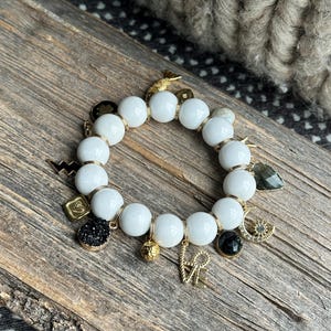May include: White beaded bracelet with gold-colored charms, including a lightning bolt, a heart, and the word "love". The bracelet also features a black stone and a gold-colored charm with a black crystal.
