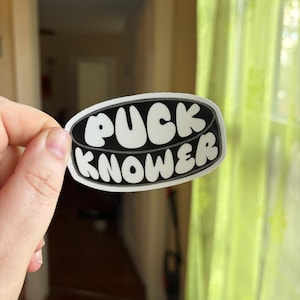 May include: An oval-shaped sticker featuring a black hockey puck design with a white border. The text PUCK KNOWER is printed in bold, white, rounded bubble letters across the center of the puck, following the curved shape of the design.