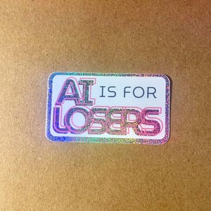 Op de afbeelding: Een rechthoekige sticker met een witte rand en de tekst "AI IS FOR LOSERS" in roze en blauwe glitterletters. De sticker heeft een holografisch effect.