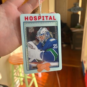 Puede incluir: Adorno azul claro con la palabra "HOSPITAL" en rojo. El adorno presenta un jugador de hockey con uniforme azul y verde. Dos curitas están en la parte inferior. Una cadena blanca está unida en la parte superior.