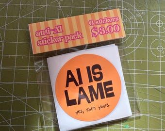 Puede incluir: Un paquete de pegatinas circulares en una funda de plástico transparente. La etiqueta superior presenta rayas naranjas y amarillas con el texto anti-AI sticker pack y 6 stickers. La pegatina principal es naranja con el texto negro AI IS LAME y yes, even yours.