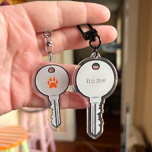 May include: Two keychains with key-shaped designs. One keychain has an orange paw print, and the other says "home" in black text. Both have silver key details and are attached to a silver and black clasp.