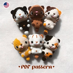 Op de afbeelding: Een set van zes gehaakte amigurumi-katten speelgoed. De katten zijn in verschillende kleuren, waaronder bruin, wit, zwart en oranje. Elke kat heeft een andere gezichtsuitdrukking en kenmerken. De katten zijn in een cirkel op een wit oppervlak gerangschikt.