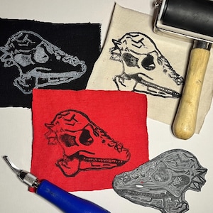 May include: A collection of dinosaur skull block prints on black, red, and cream fabric. A linoleum block and a roller are also visible. The prints are in black ink, and the linoleum block is gray. The tools suggest a printmaking theme.